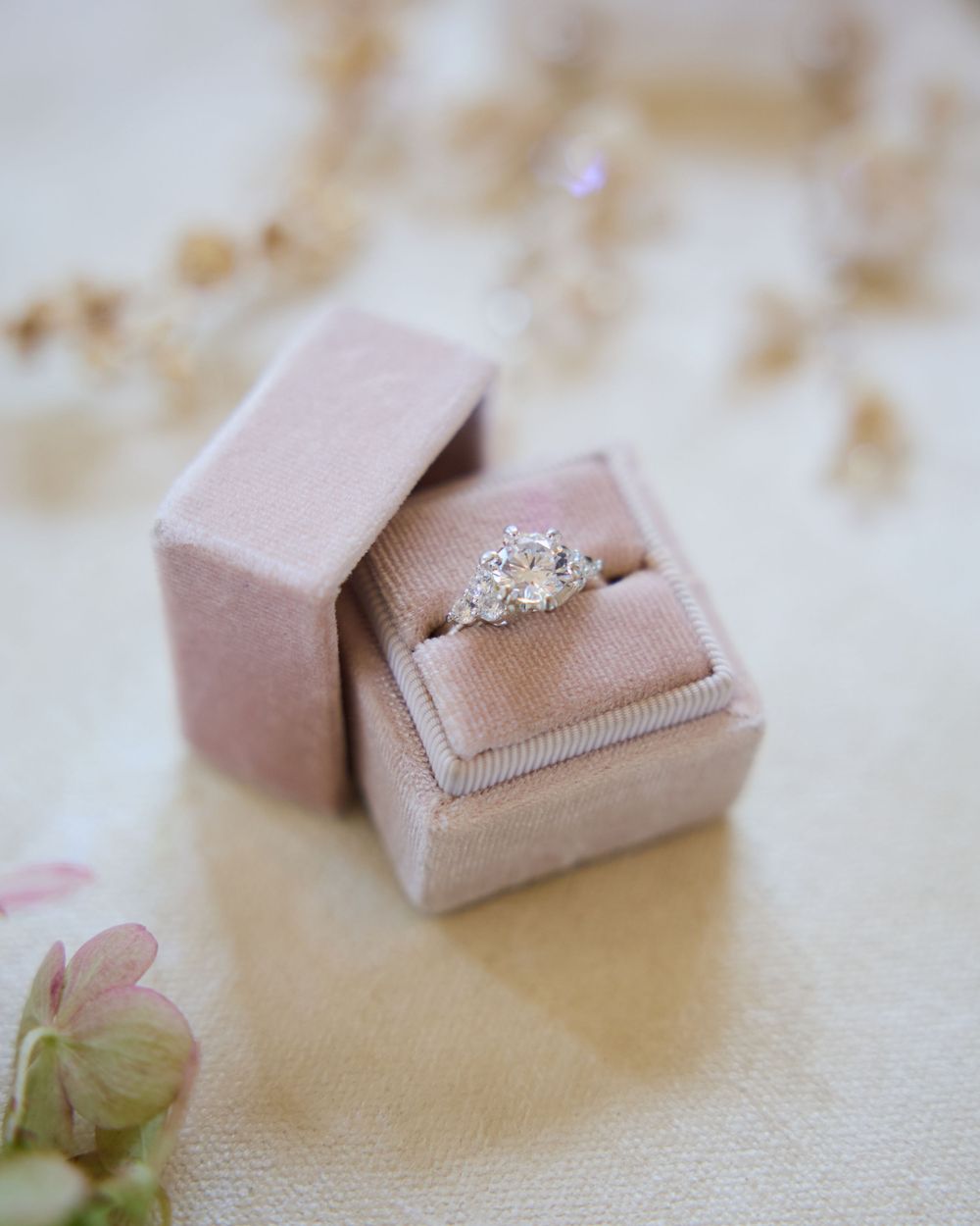 A sparkling diamond engagement ring sits in a blush pink velvet ring box with a small green flower beside it.
