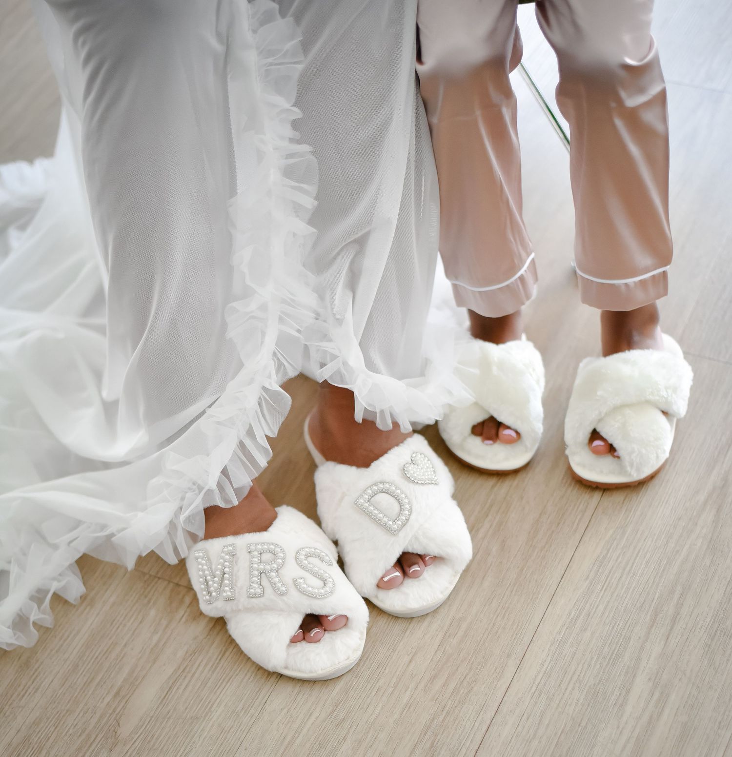 Matching white bridal slippers with 'Mrs' text on wooden floor next to wedding dress hem.