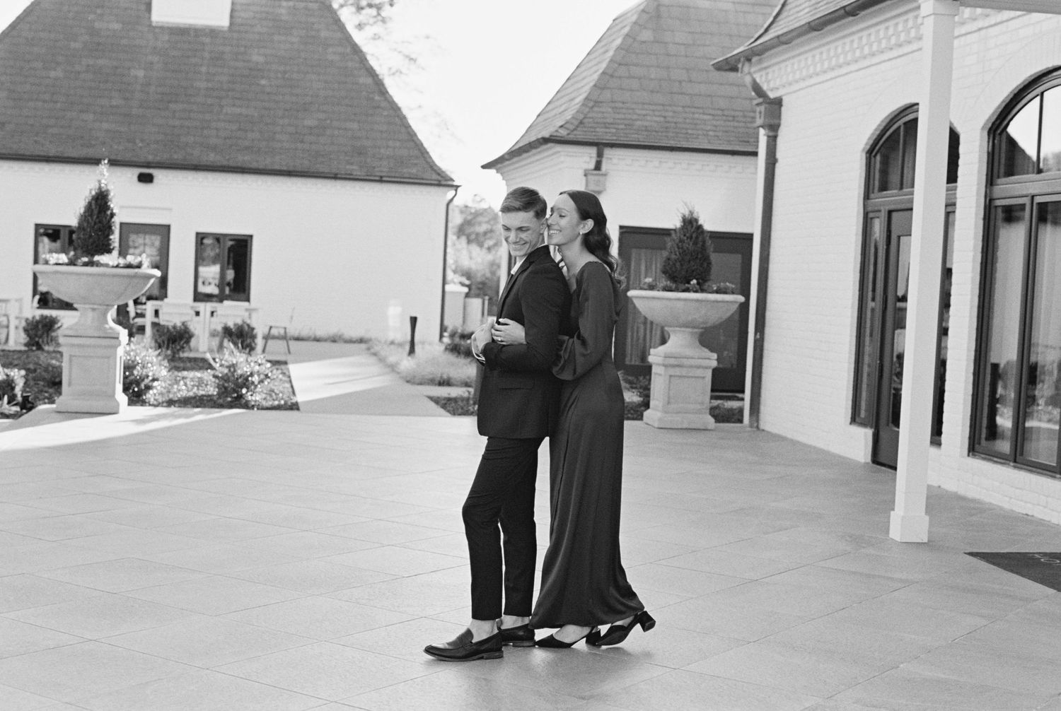 A couple embraces while dancing together on a patio in front of white buildings with arched doorways.