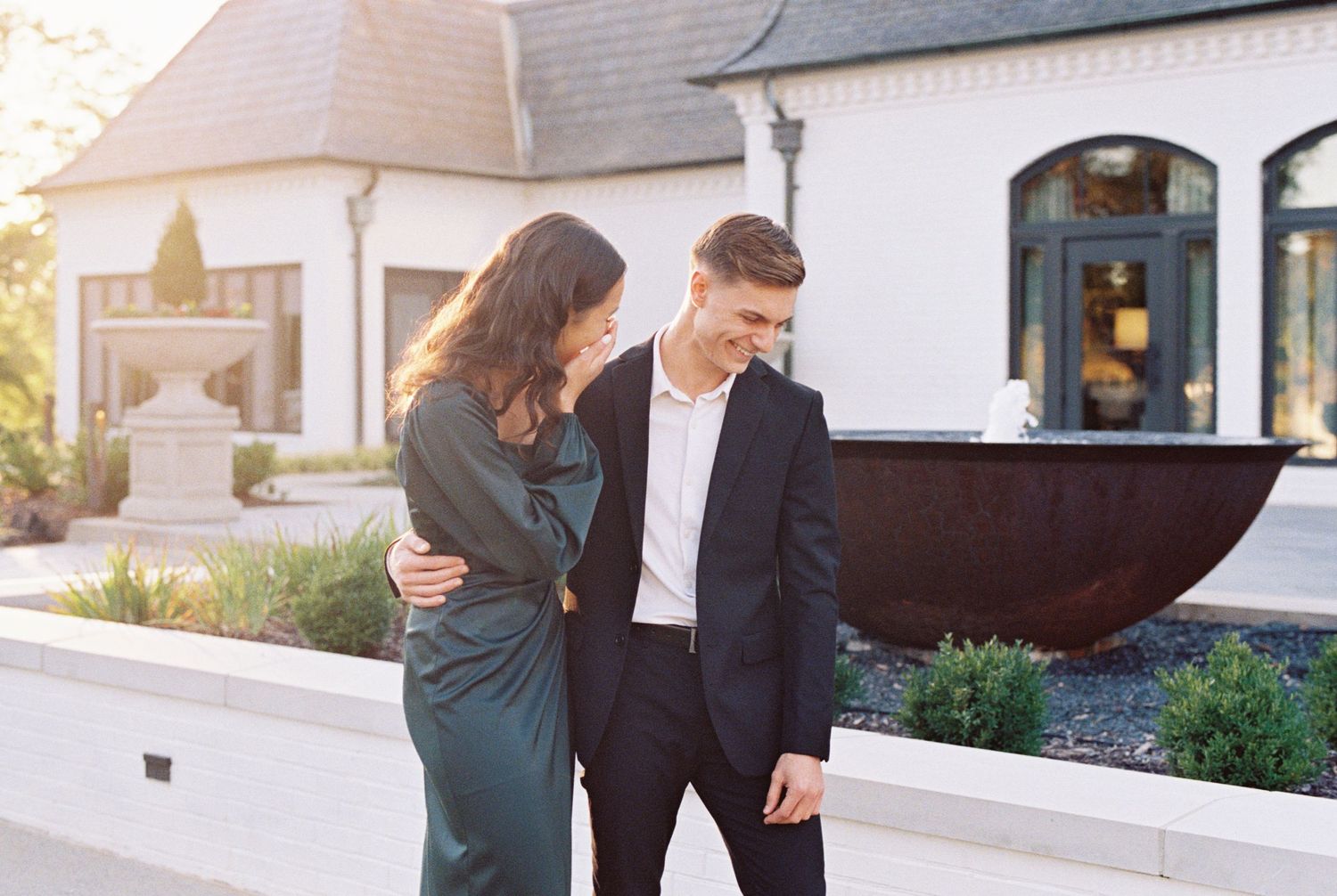 Romantic couple in elegant attire pose together near white buildings and landscaping.