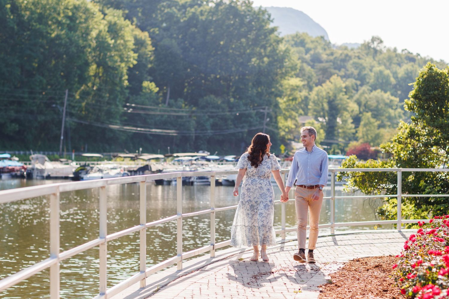 Two people stroll along a lakeside path in the sunlight, with boats docked in the marina behind them.