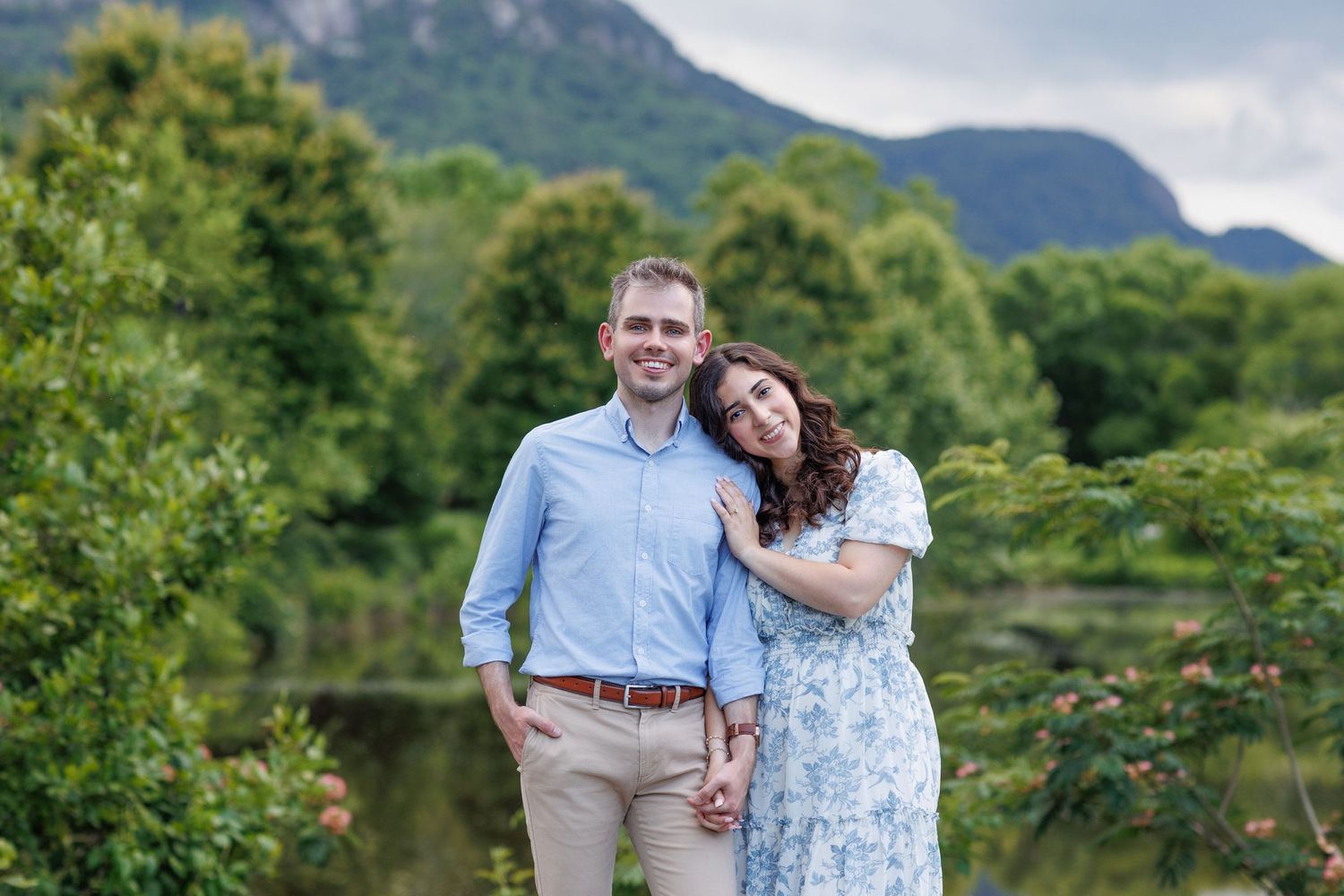 Couple embracing in a scenic mountain landscape with lush greenery and water.