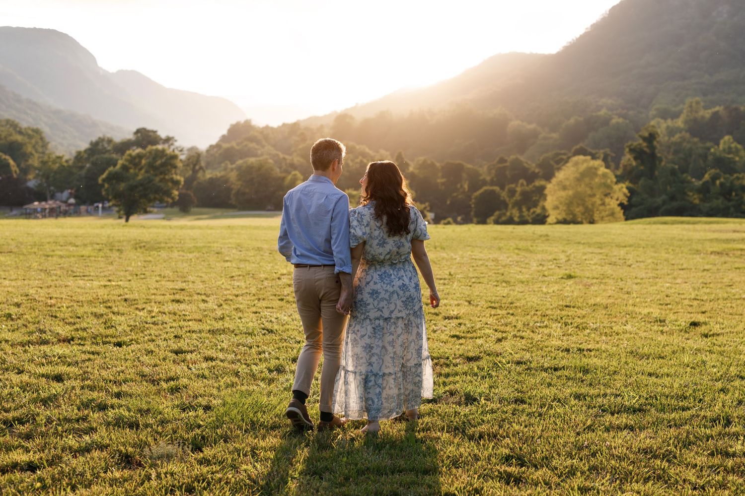 A couple walks hand in hand through a sun-drenched meadow with mountains in the background at golden hour.