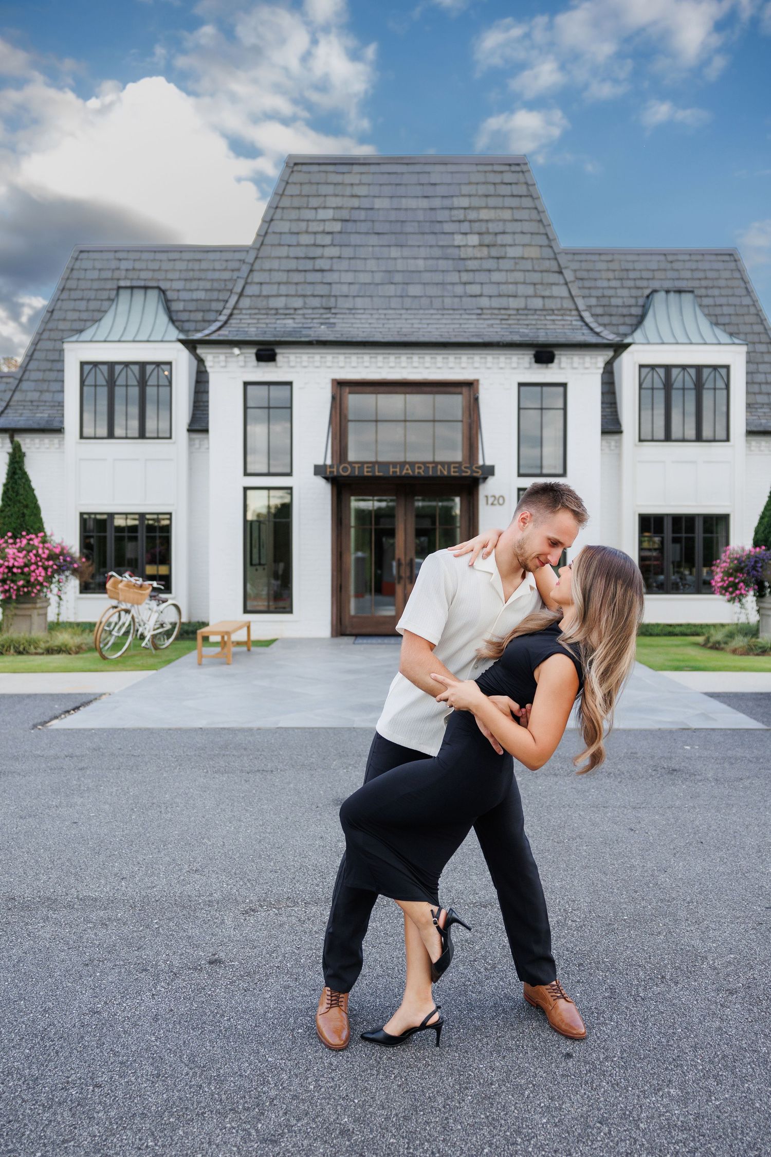 A romantic couple embraces in a dance pose in front of a modern white mansion with black trim and a striking facade.