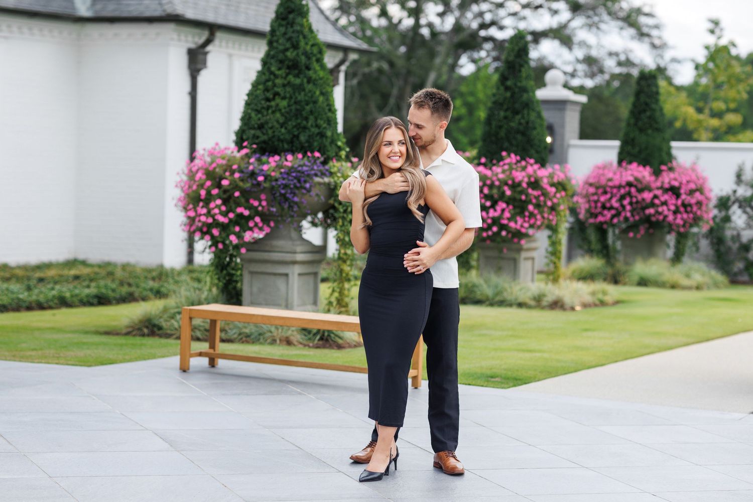 A couple embraces on a patio with pink flowers and greenery in the background.