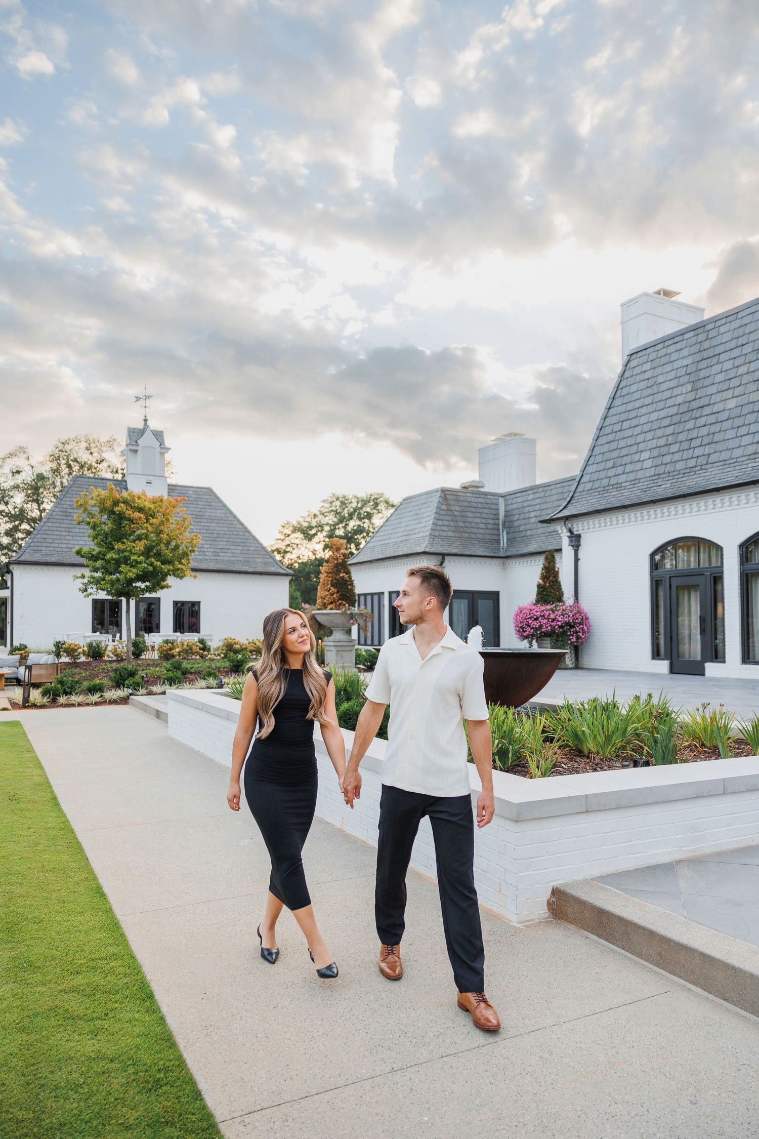 A couple walking hand in hand on a path at sunset in front of white modern farmhouse style buildings.