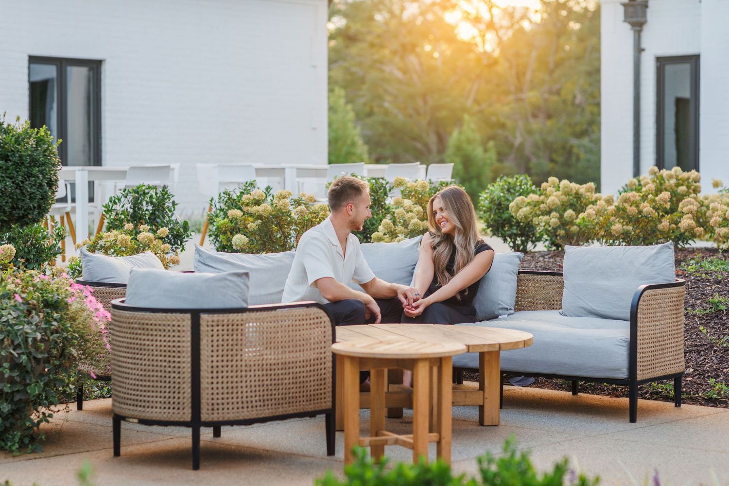 Couple relaxes on elegant patio furniture at sunset with lush greenery and white exterior walls in background.