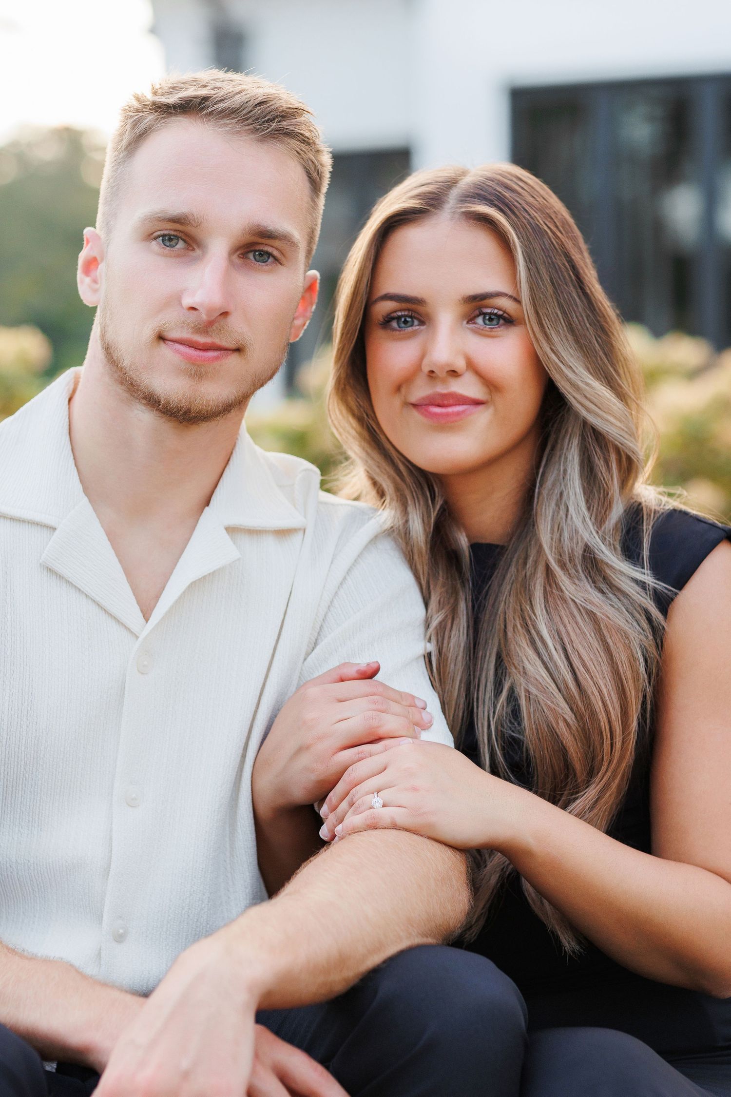 A couple in white and black attire pose together outdoors in a romantic engagement-style portrait.