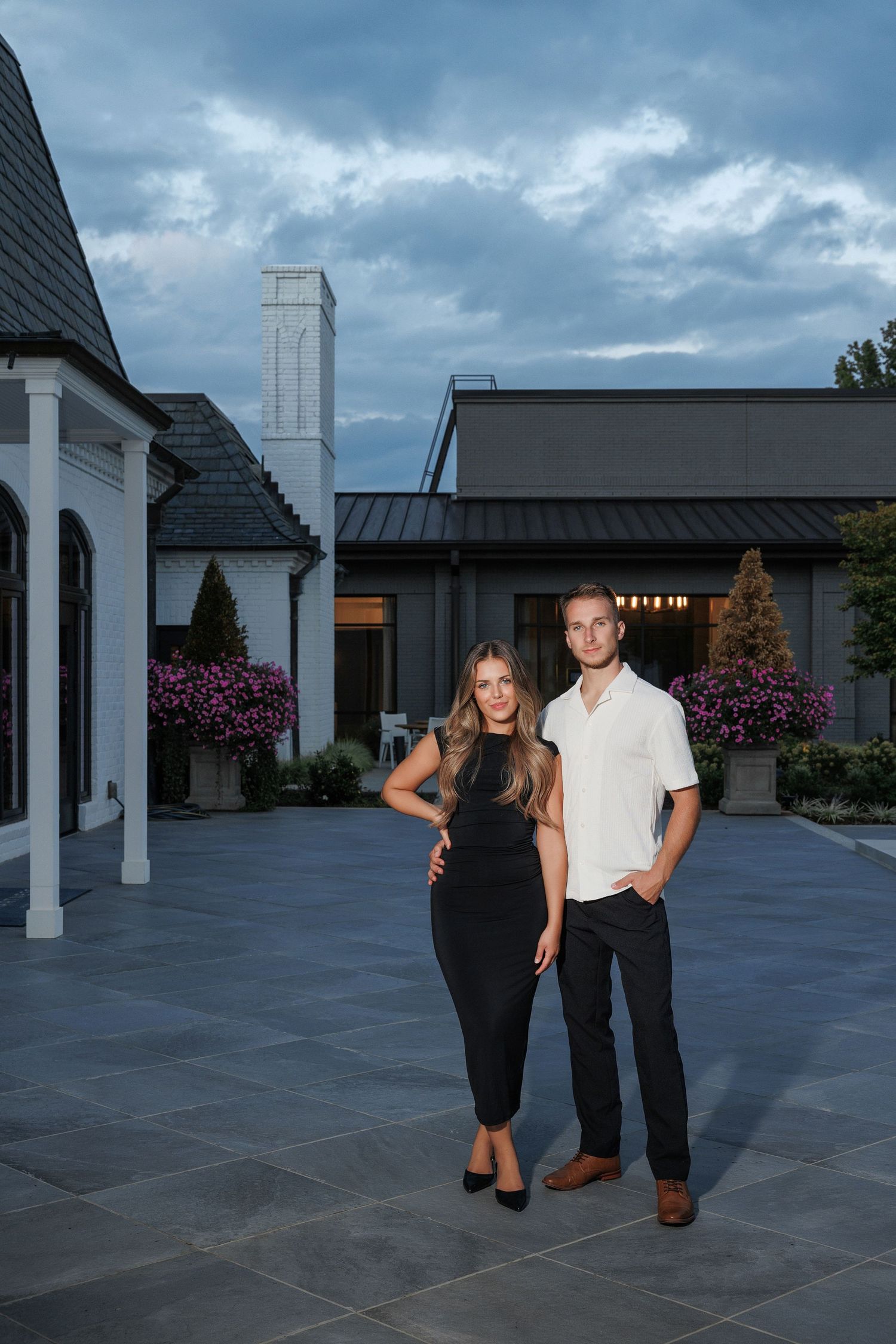 A couple stands in an elegant courtyard at dusk with modern architecture and flowering plants in the background.
