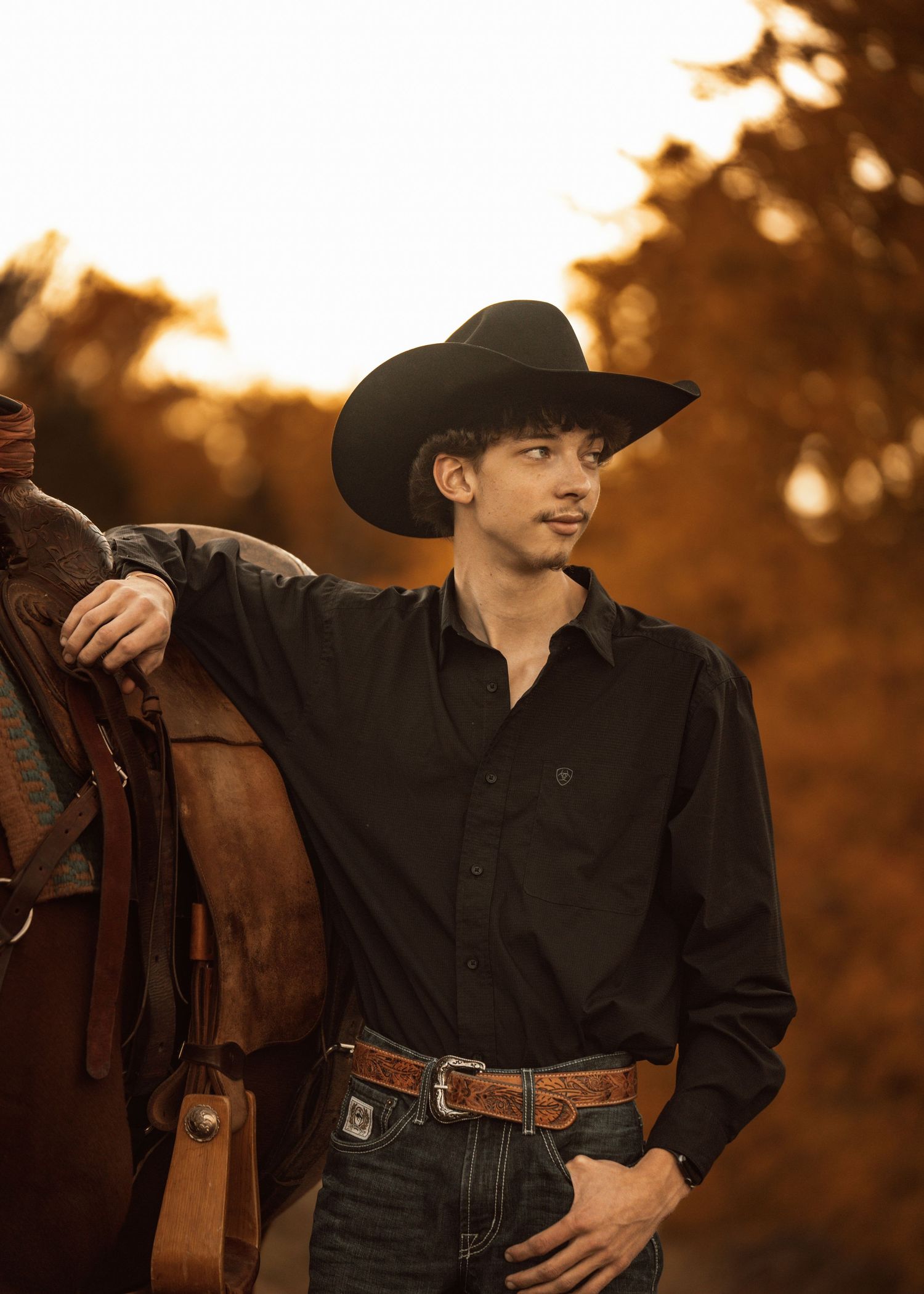 Person in western attire poses against sunset with leather saddle.