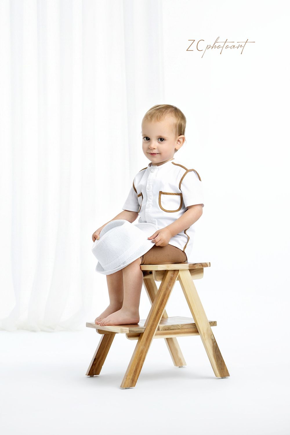 Adorable toddler in white outfit sitting on wooden step stool holding a hat against white backdrop.