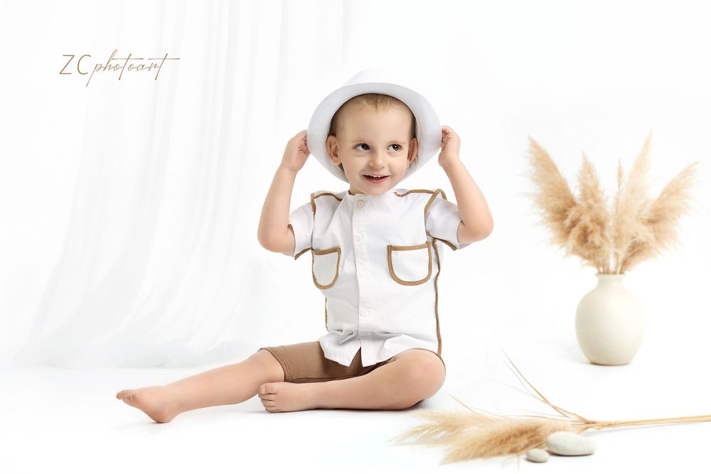 Smiling toddler in white hat and outfit sits on floor with pampas grass decor in bright studio photo.