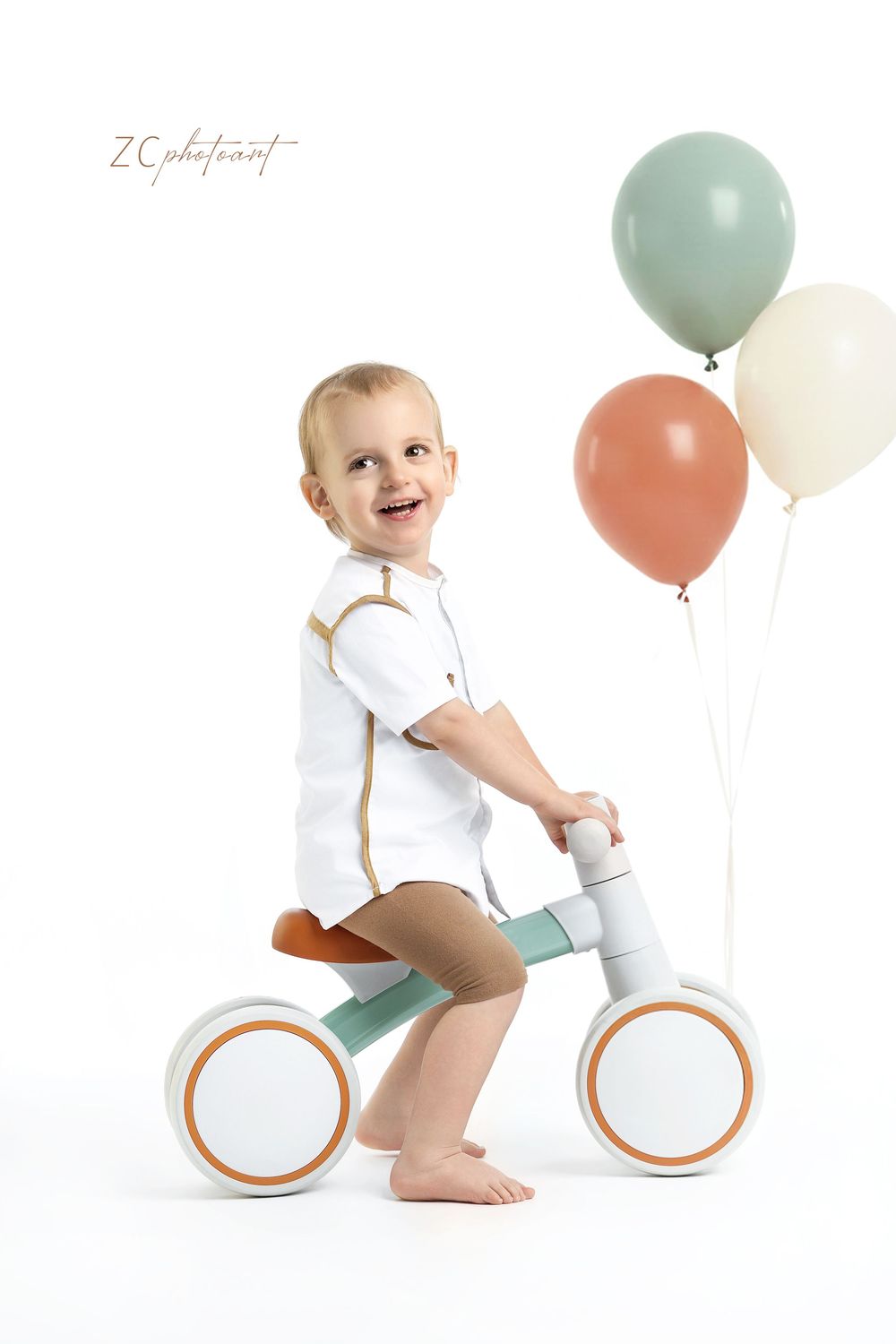 Smiling toddler boy riding a balance bike with colorful balloons on white background.