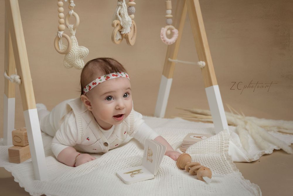 A baby in white clothing plays on a beige carpeted floor with wooden toys and accessories.