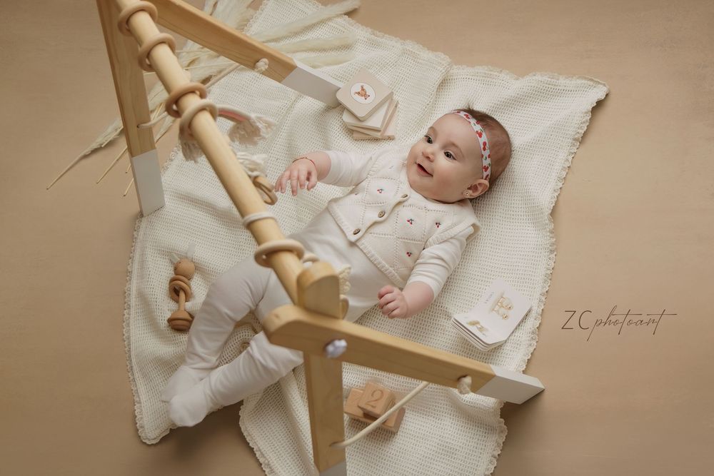 A baby lies peacefully on white bedding next to a wooden spindle in a warm, natural light setting.