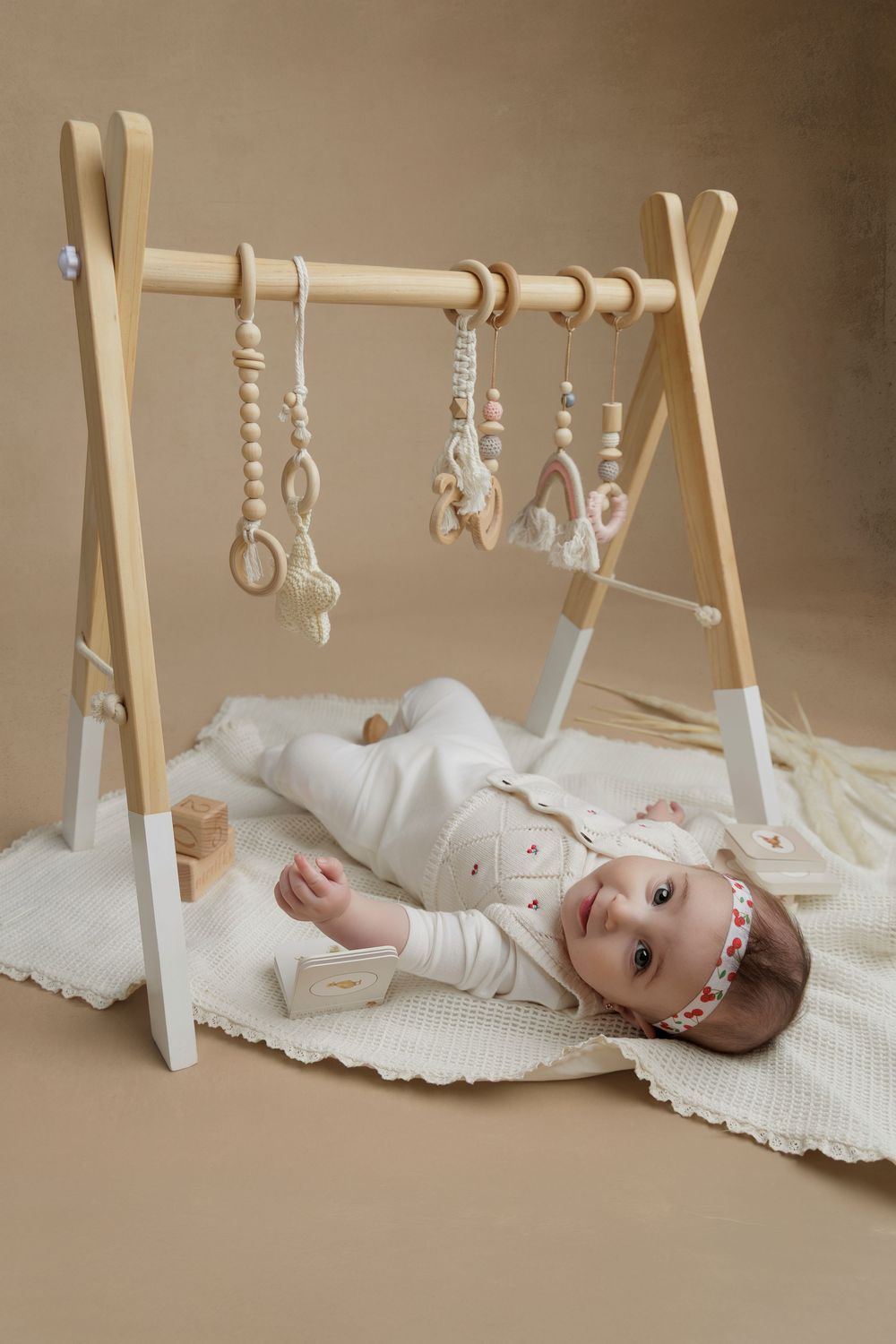 Baby lies on cream blanket under wooden activity gym with hanging toys in natural light studio setting.