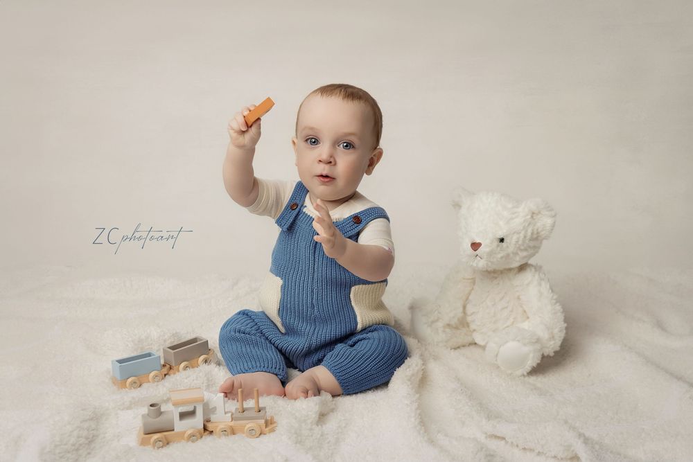 Baby in blue overalls sits on white backdrop with stuffed bunny toy and wooden blocks.