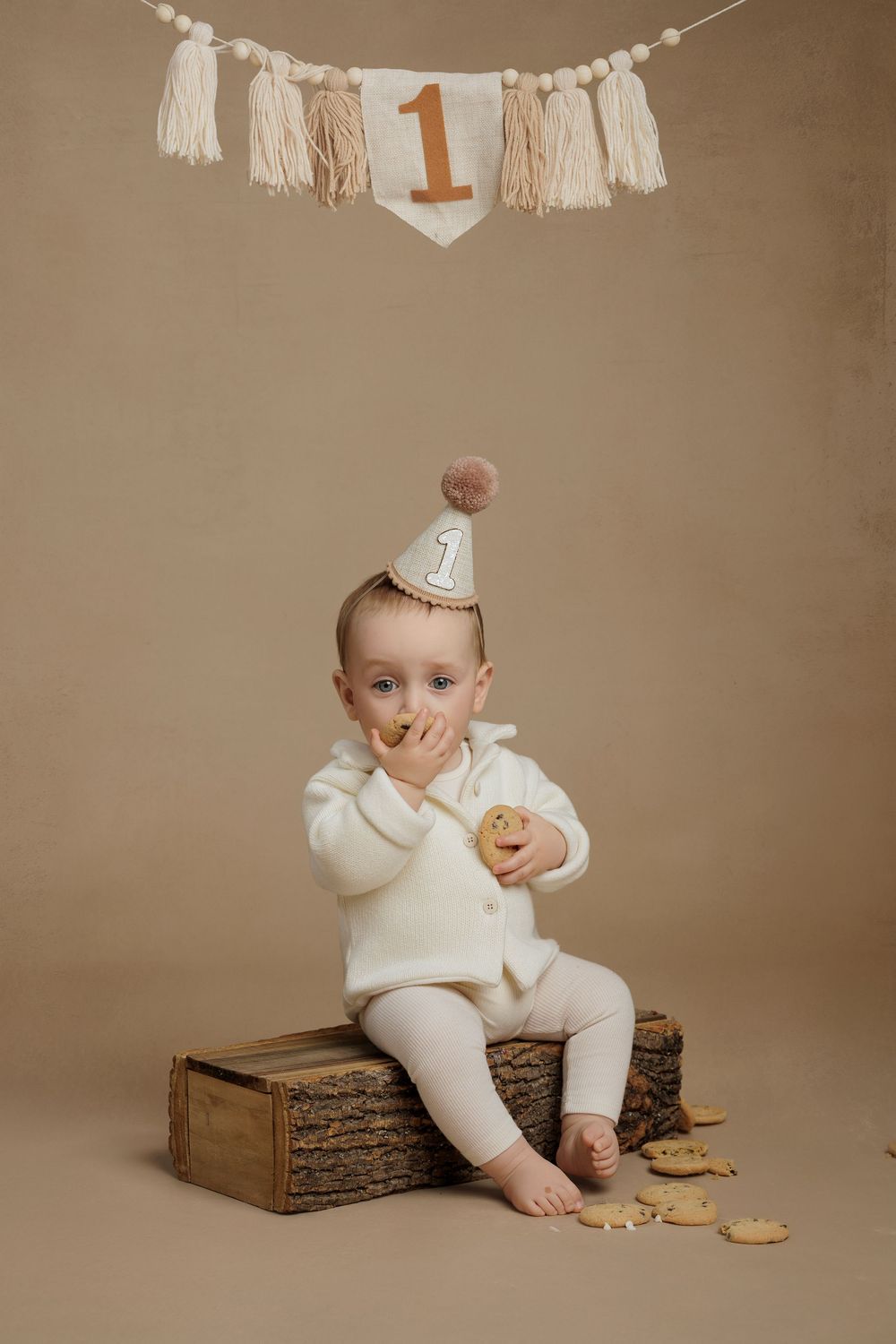 A baby in a white outfit sits next to a wooden box with decorative number one banner and tassels in a studio setting.