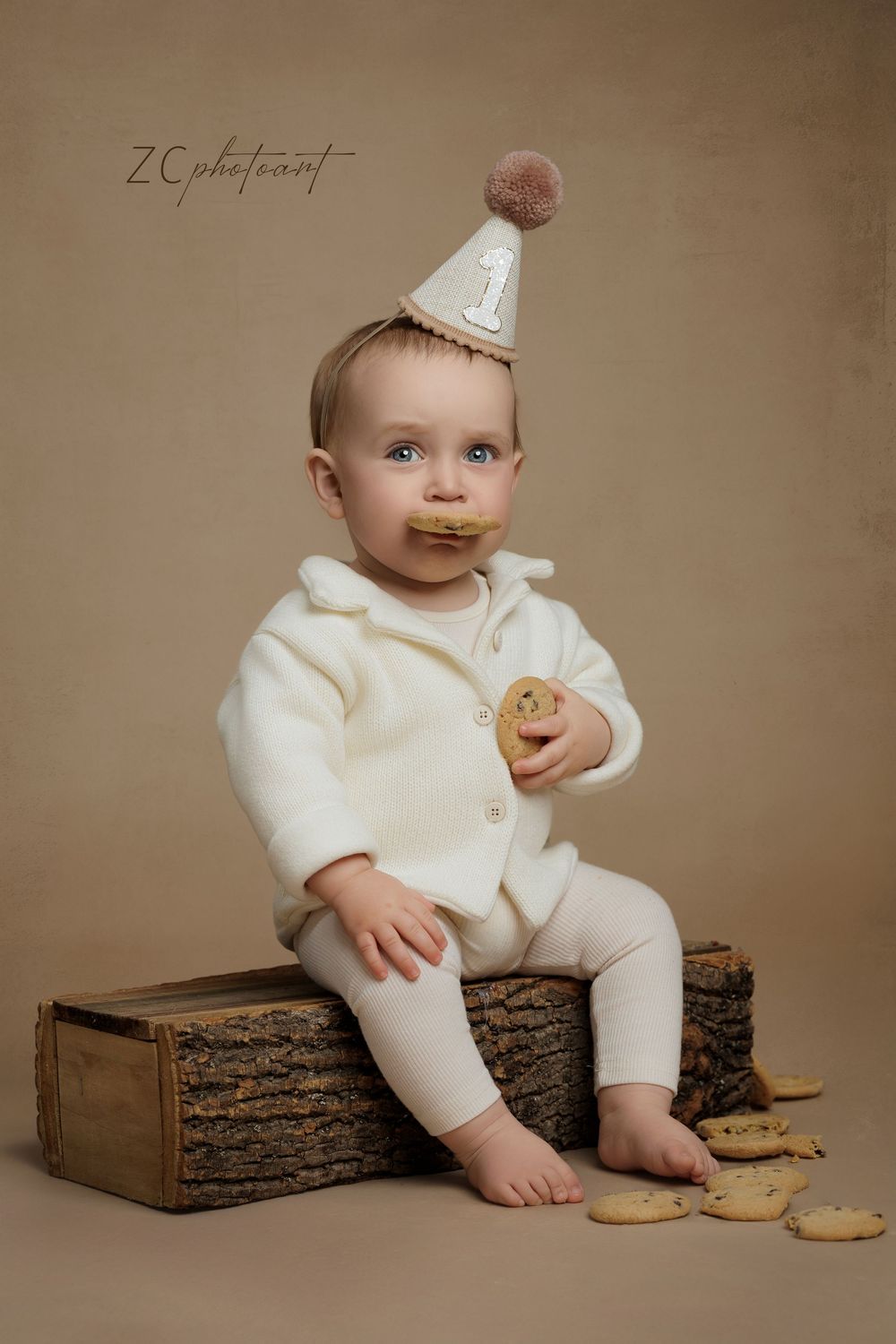 Small child in white outfit and party hat sits on rustic wooden box with cookies, creating charming first birthday scene.