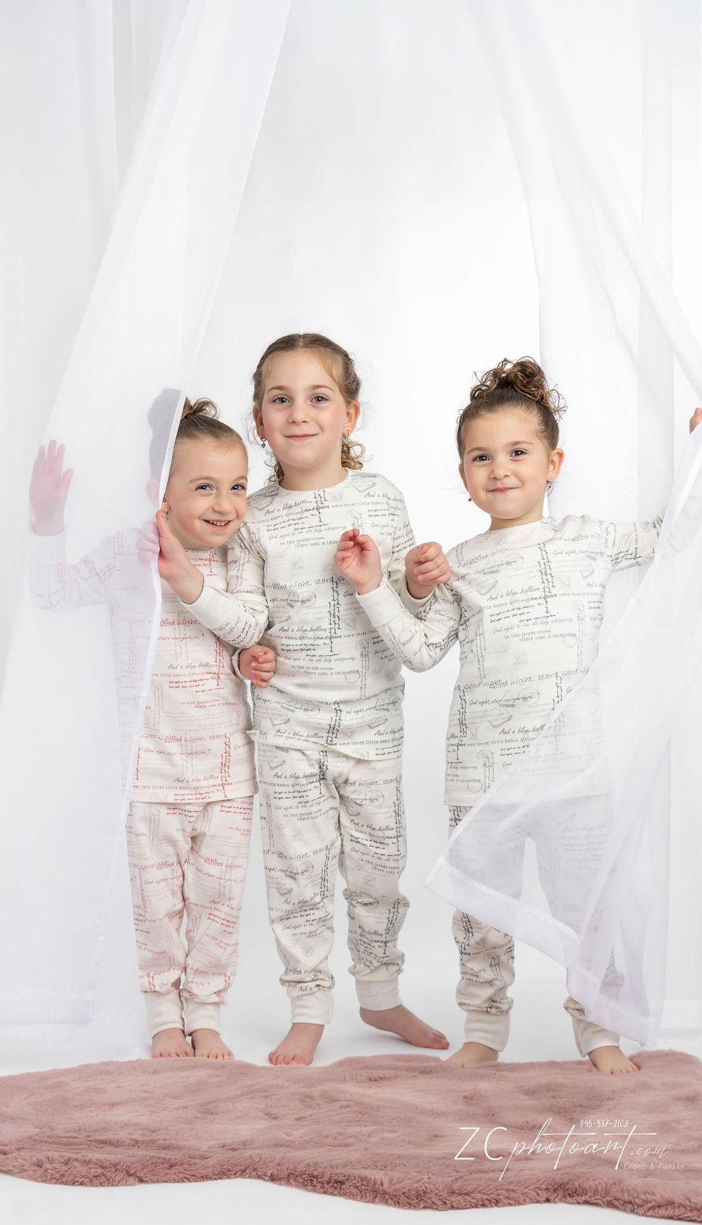 Group of young children wearing matching cream and text-patterned outfits pose together against white curtains and pink carpet.