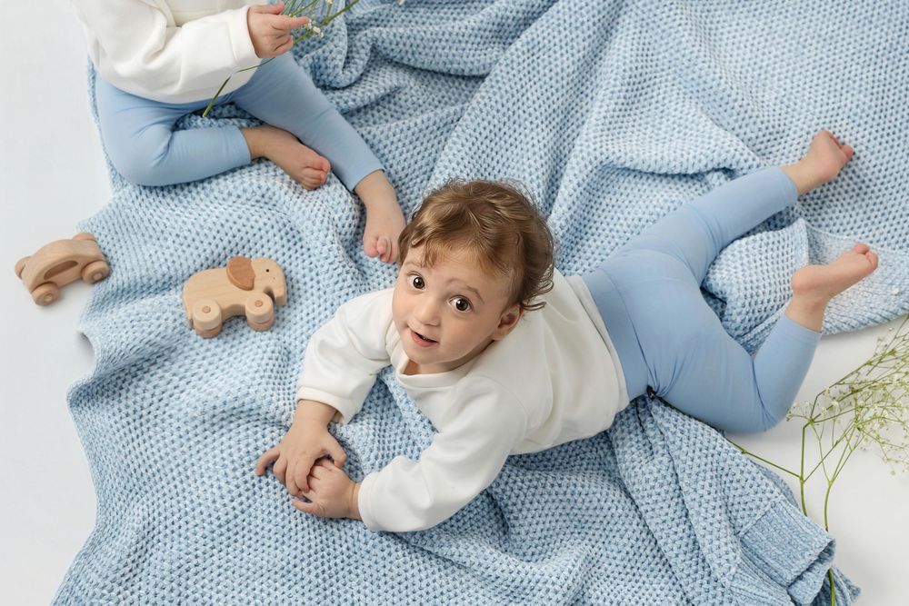 Baby in white onesie lying on blue knit blanket playing with wooden toy blocks and teethers.