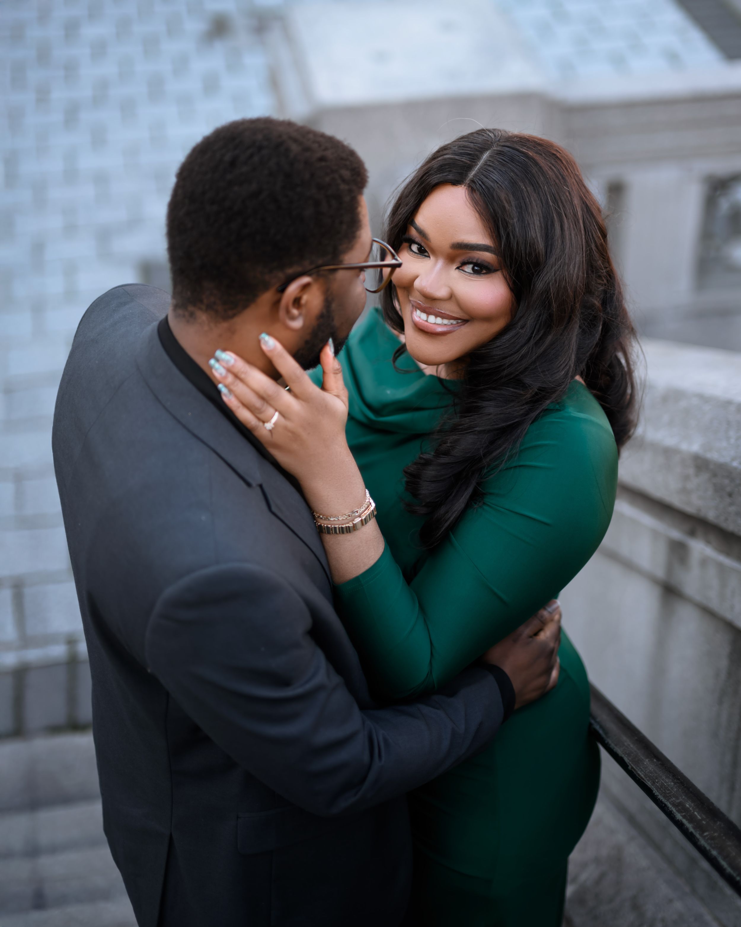 A romantic couple embraces on steps, with the person in emerald green showing off an engagement ring.
