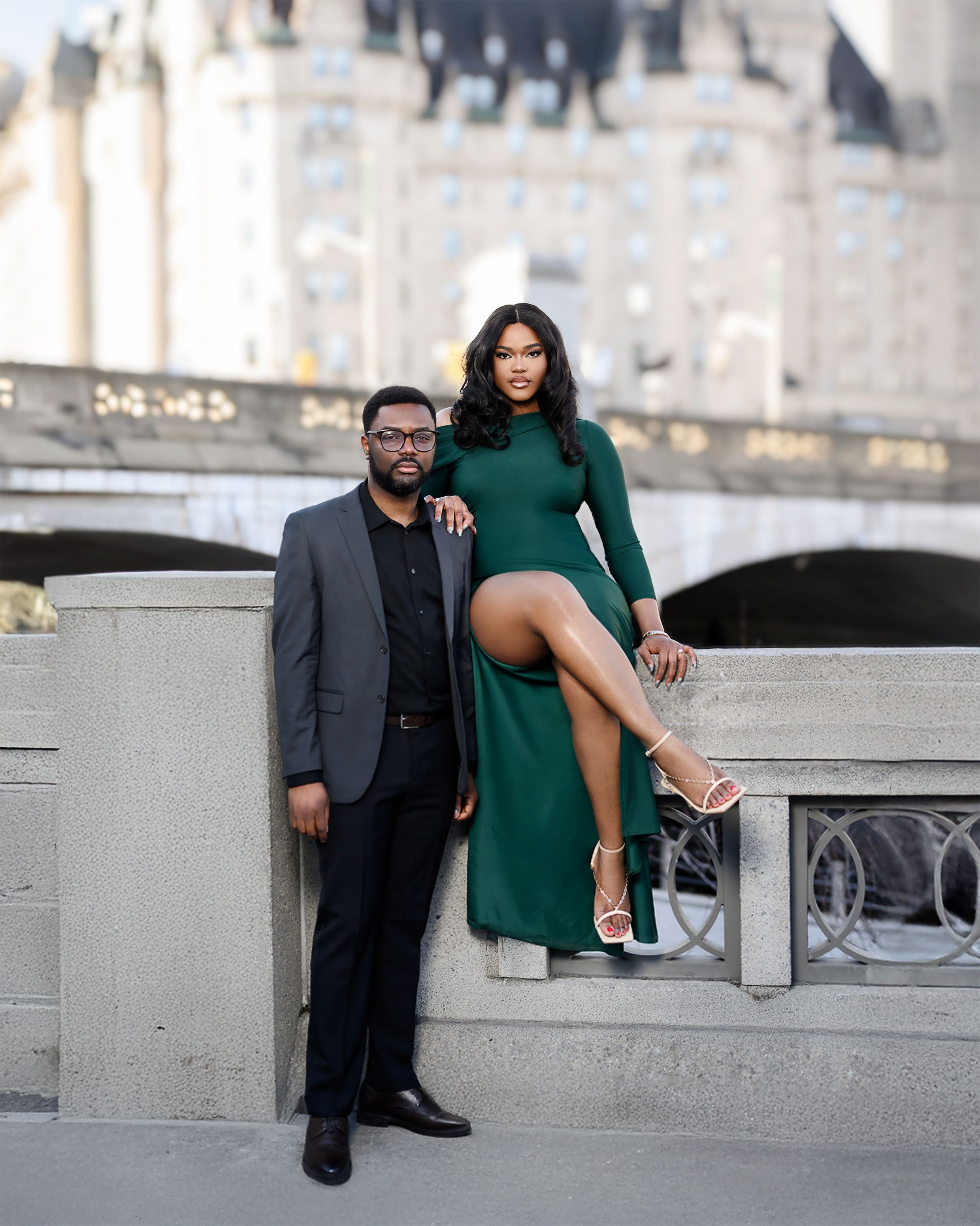 A couple poses elegantly in front of a castle backdrop, with the woman wearing a green dress seated on a stone wall.