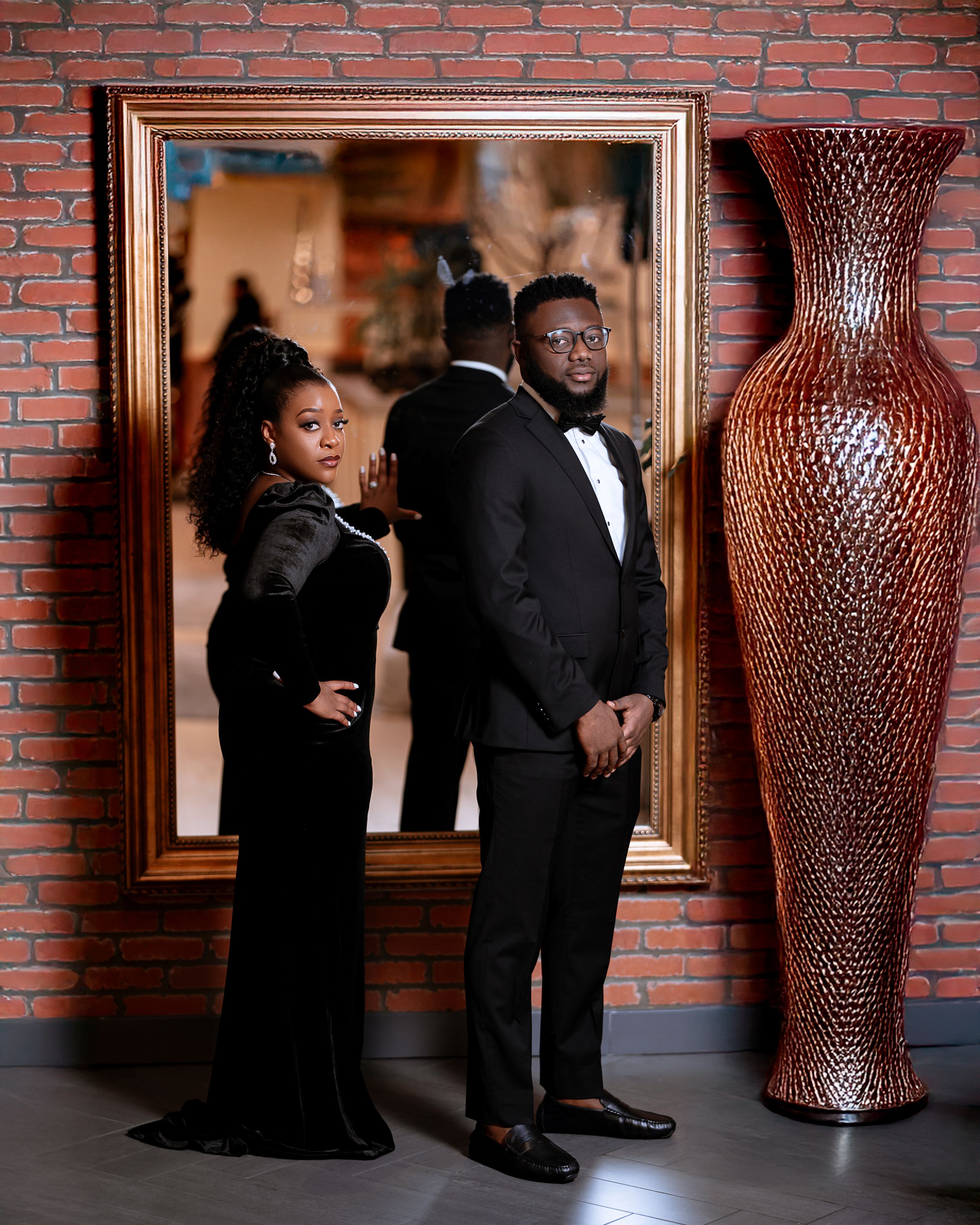 Elegant couple in black formal attire pose for glamorous photo against brick wall with decorative tall vase.