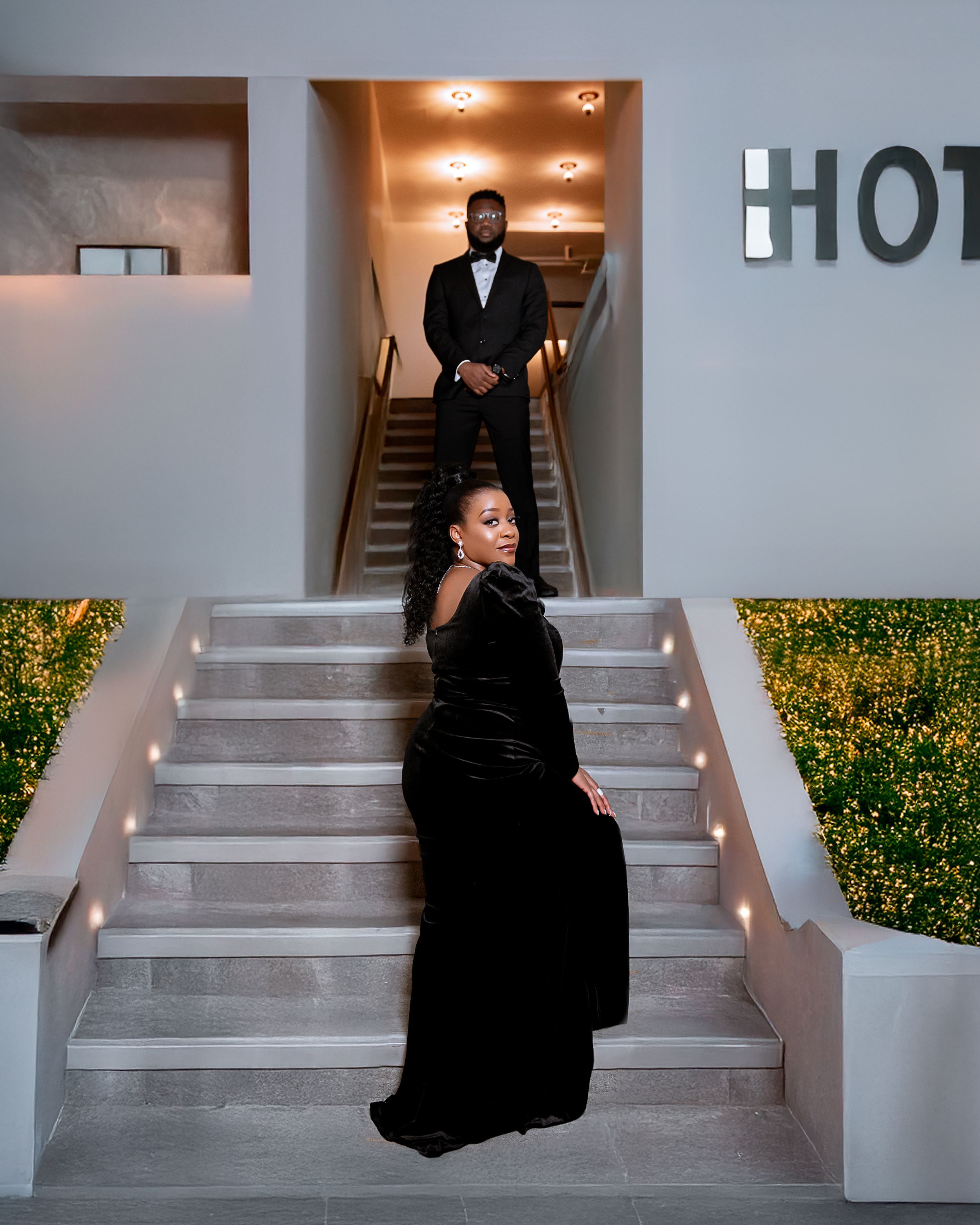 Elegant couple dressed in formal black attire pose dramatically on illuminated white hotel steps at night.
