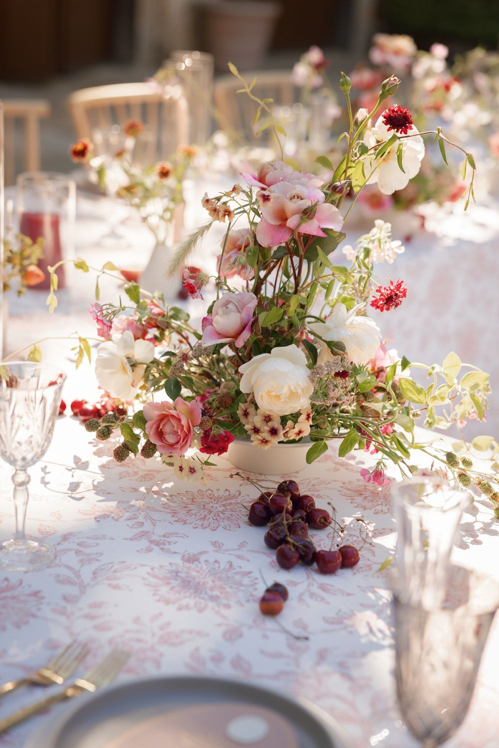 Elegant table setting with pink and white floral centerpiece, crystal glasses and scattered berries on lace tablecloth.
