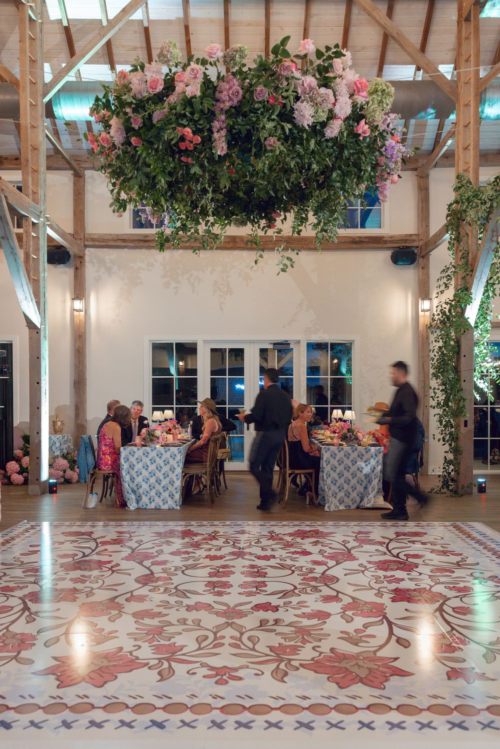 Elegant reception venue with patterned floor, exposed wooden beams, hanging floral arrangements and guests seated at tables.