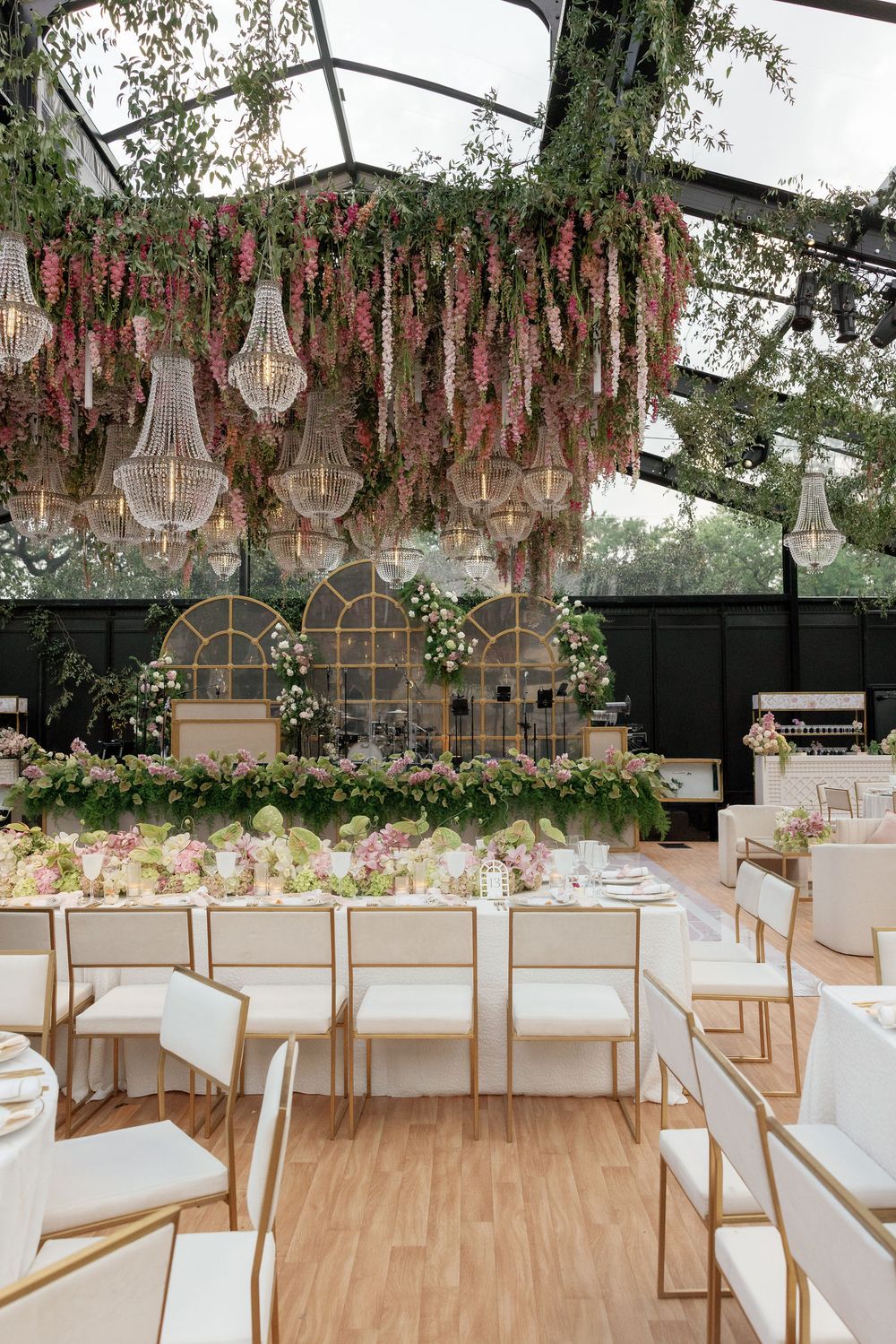 Elegant wedding reception tables under hanging crystal chandeliers and pink floral arrangements in a greenhouse setting.