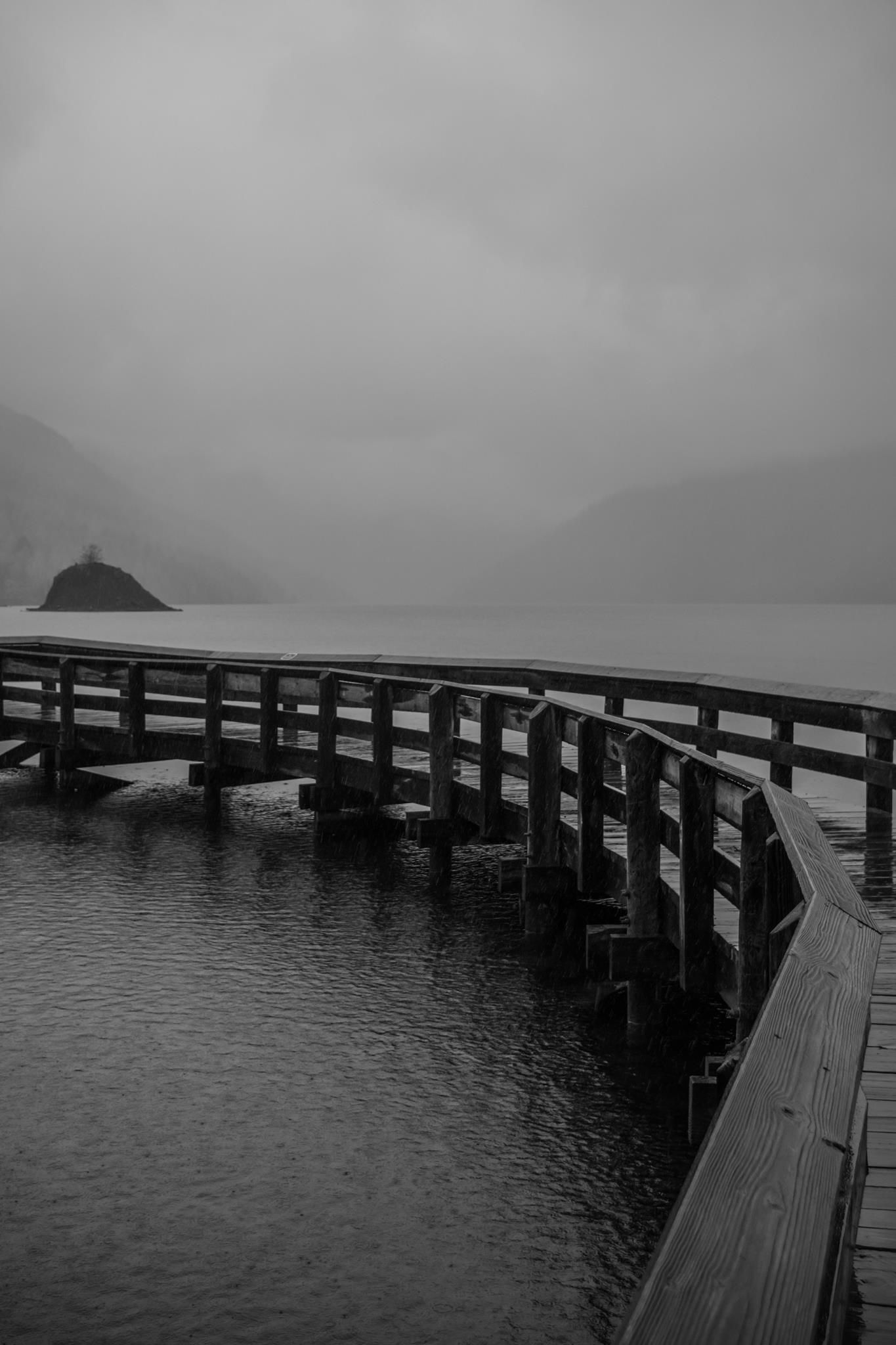 Wooden pier extends into a misty lake surrounded by mountains on a foggy overcast day.