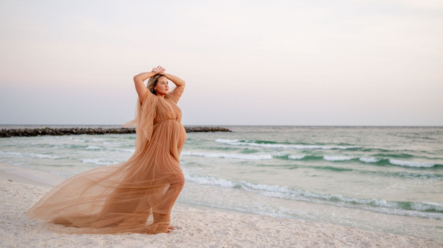 Romantic beach maternity portrait in flowing peach dress with ocean waves at sunset.