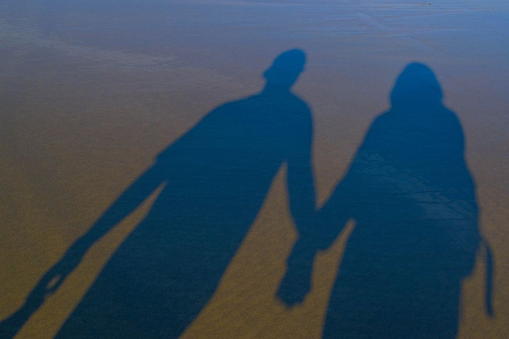 Two elongated shadows hold hands on a sandy beach at sunset.