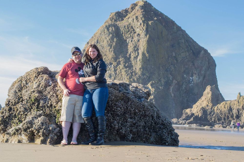 Two visitors pose at Haystack Rock on Cannon Beach, Oregon with the iconic rock formation looming in the background.