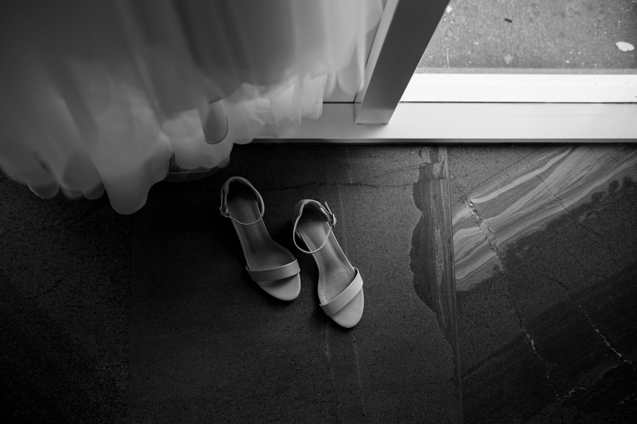 Wedding shoes placed beneath flowing white fabric near a window in black and white.