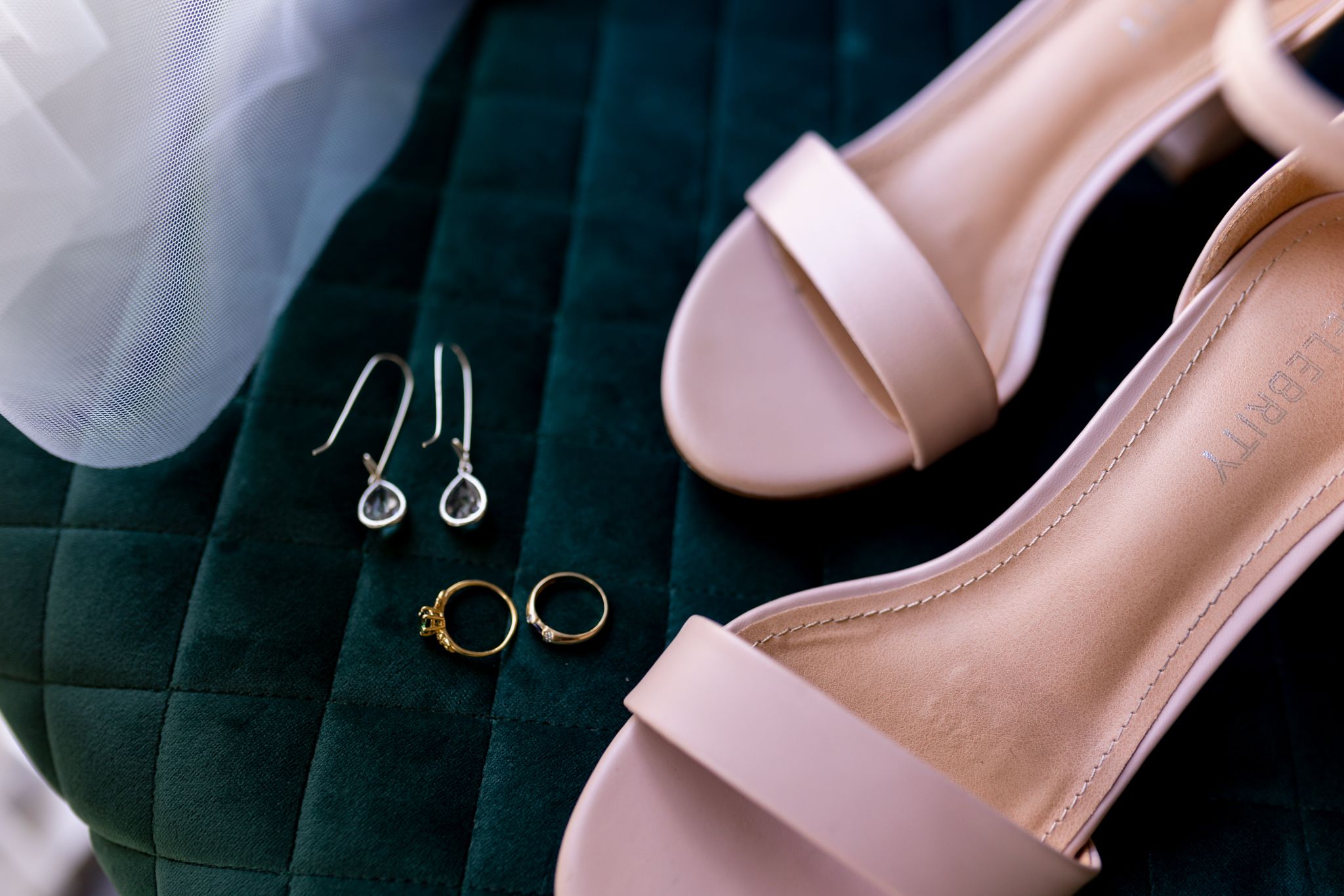 Wedding accessories including pink sandals, earrings and rings arranged on dark velvet fabric.