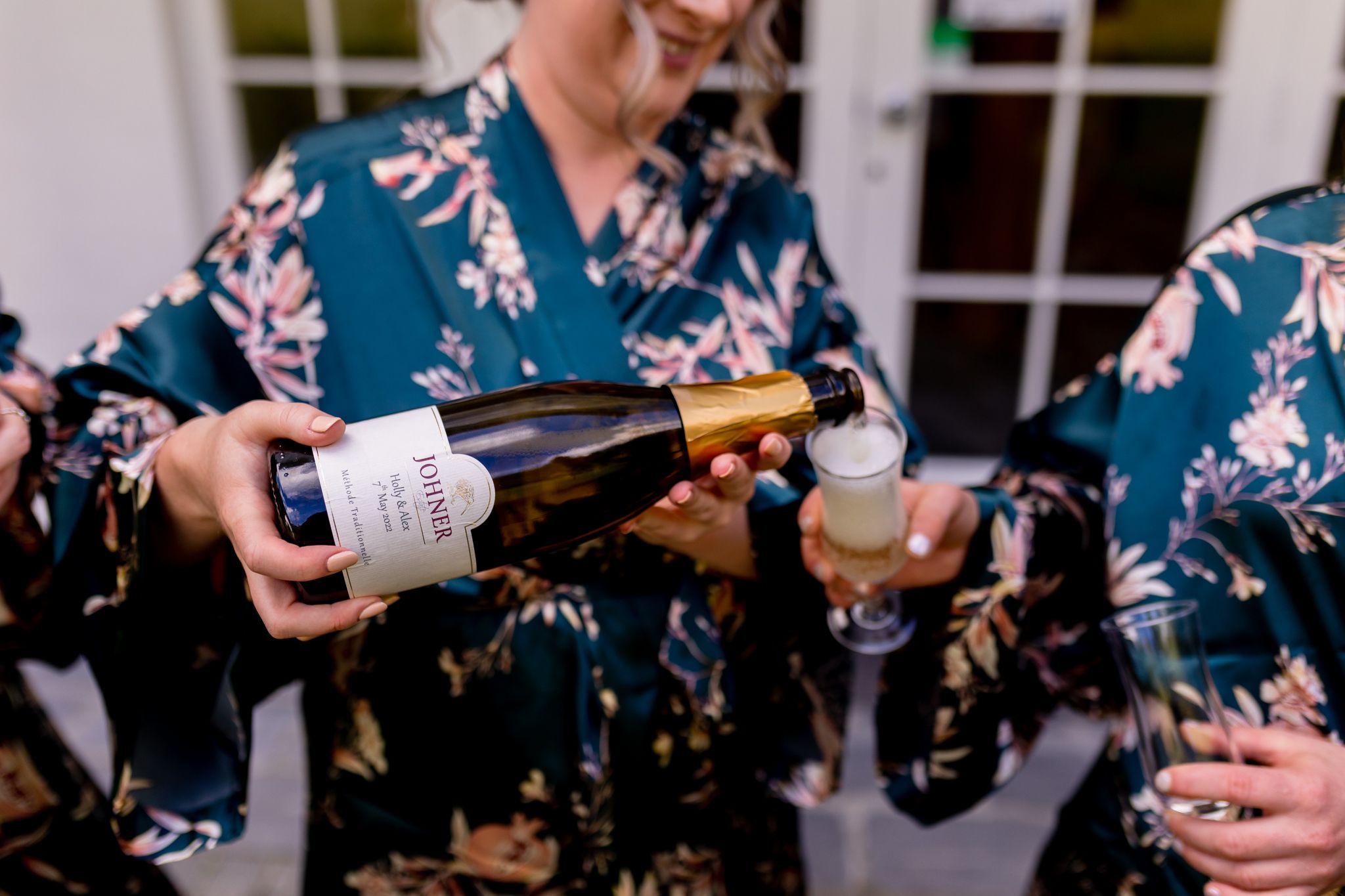Person pouring champagne while wearing a blue floral robe during pre-wedding preparations.