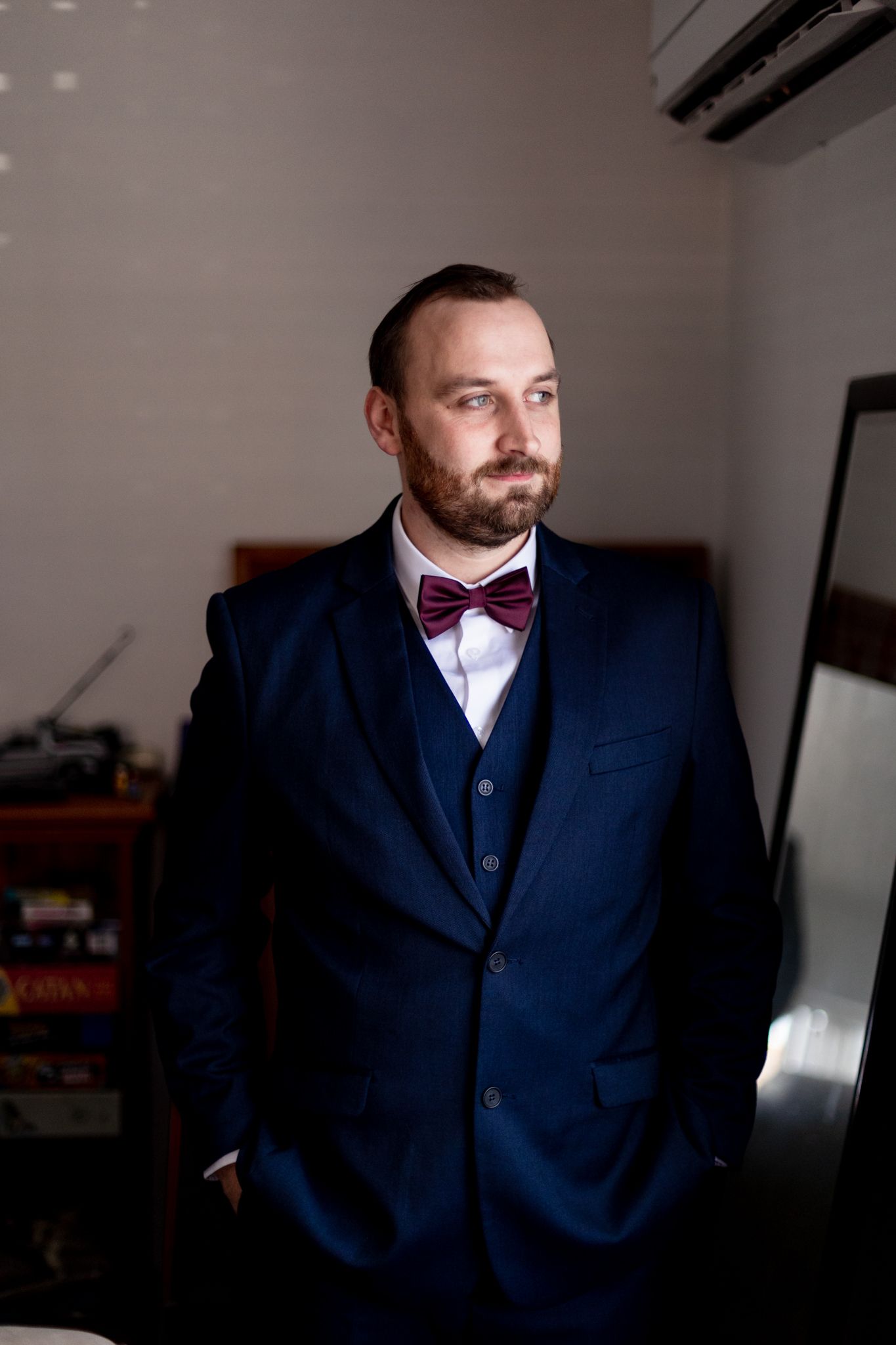 Groom in navy three-piece suit and burgundy bow tie poses in natural light.