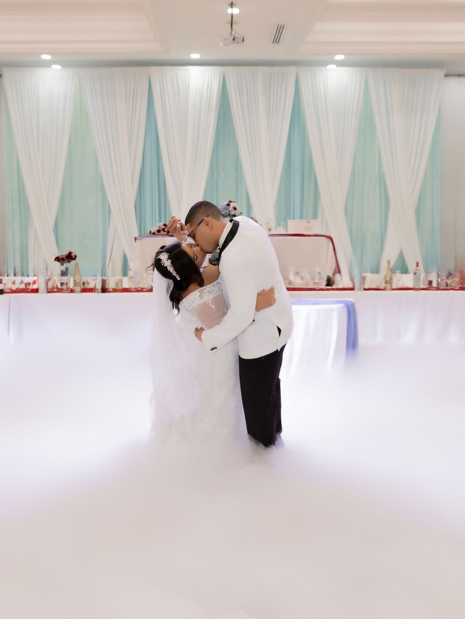 Wedding couple sharing intimate kiss on dance floor with teal and white backdrop.