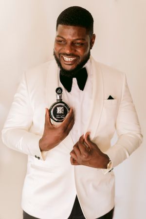 Stylish groom in white tuxedo jacket and bow tie smiling while holding luxury perfume bottle.