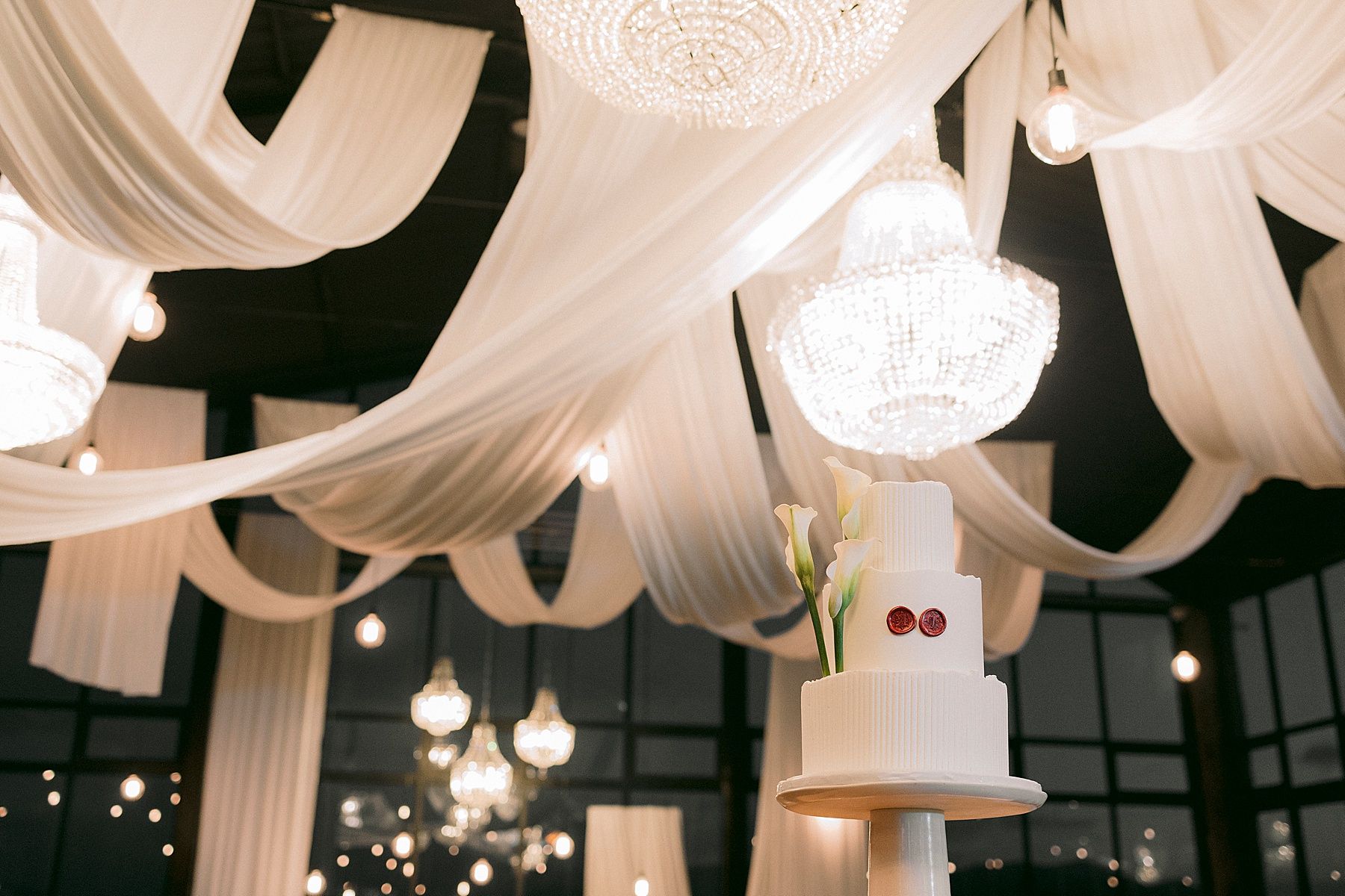 Elegant wedding reception venue with white draped ceiling, crystal chandeliers, and a two-tier white wedding cake. Bakenhof, Wellington.