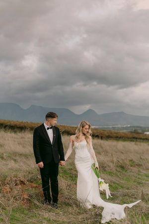 Bride and groom holding hands in a golden field with dramatic cloudy skies and mountain range backdrop. Bakenhof, Wellington.