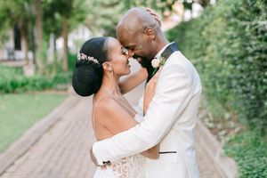 Black bride and groom in white tuxedo share romantic forehead touch on brick pathway surrounded by lush greenery. Fusion Boutique Hotel.