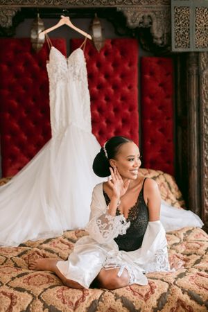 Beautiful Black bride in lace robe sits on ornate bed before beaded mermaid wedding gown on hanger.