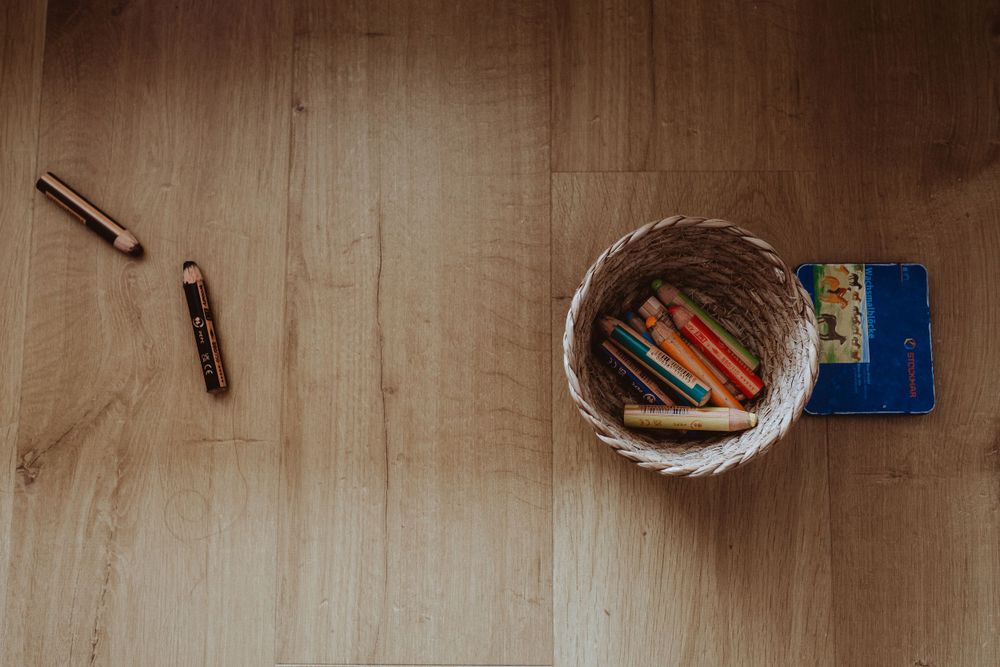 Overhead view of wooden basket containing colorful art supplies on hardwood floor.
