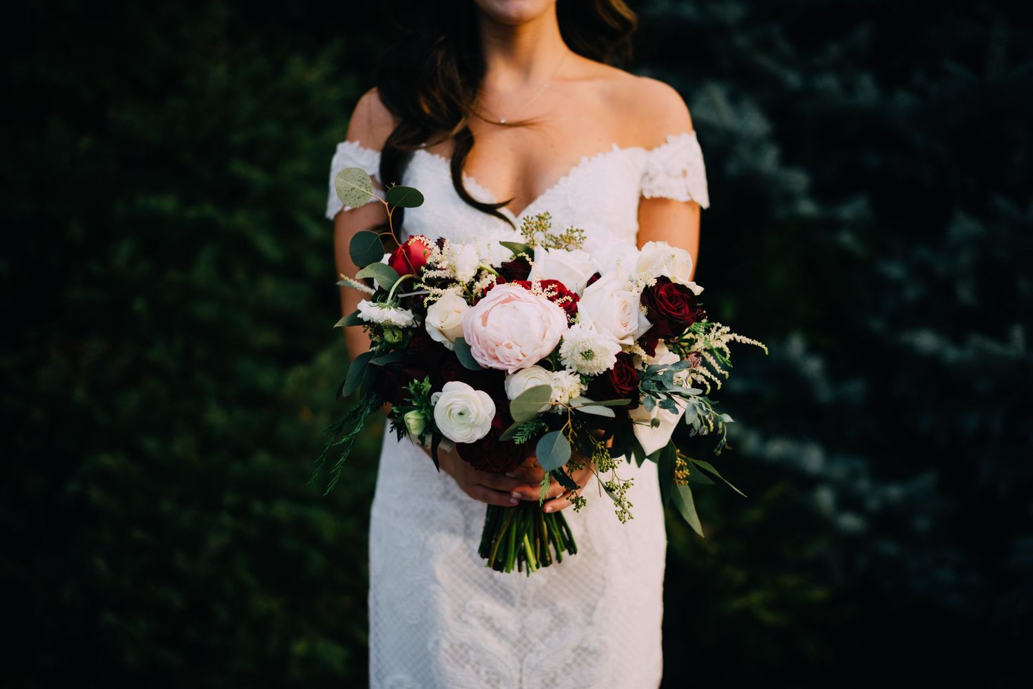 Megan is holding a gorgeous bouquet filled with peony's, roses, green leaves. Her dress hangs off of her shoulders.