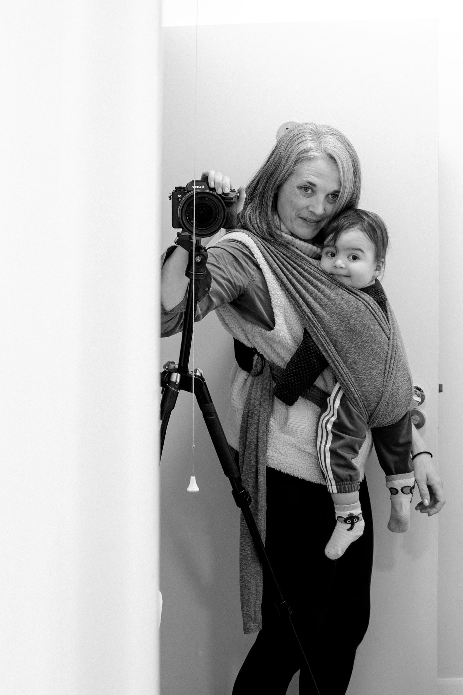 Black and white photo of photographer with camera on tripod carrying baby in grey wrap carrier.