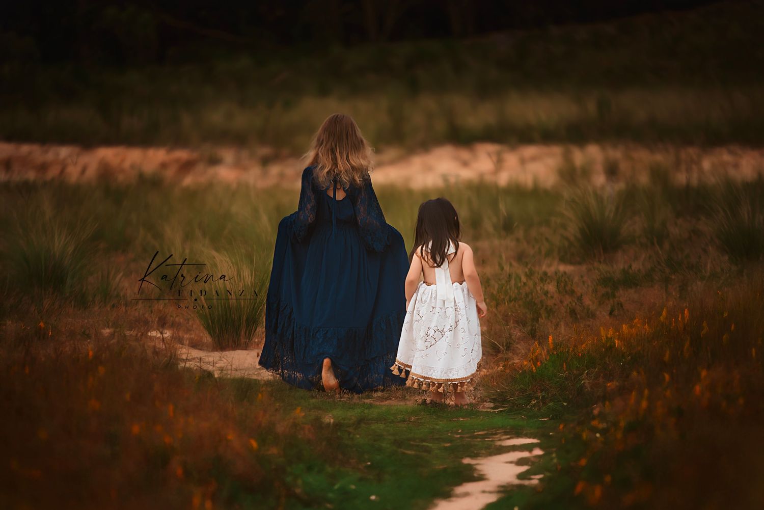 Two silhouettes walk along a winding dirt path through a moody autumn field at sunset.
